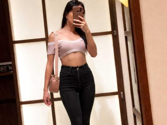 PamelaKander with long black hair, wearing a pink off-shoulder top, black high-waisted jeans, pink crossbody bag, and holding a phone, showing her small tits and slim ass.