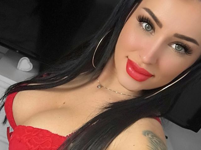 LovelyMichellex69 with red lipstick, hoop earrings, and red lace bra, showing her tits and tattooed arm, smiling at the camera.