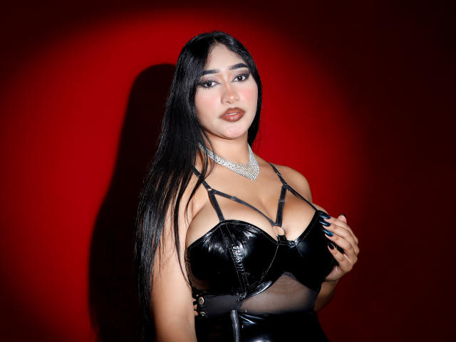 MelisaJackie with long black hair, wearing a shiny black leather top that highlights her big tits, and a diamond necklace, against a red background, looking confidently at the camera.