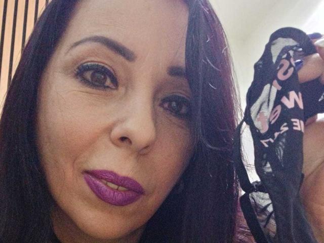 LauraSin with dark brown hair, purple lipstick, holding black lace panties with pink letters, looking into camera, slightly smiling, showing her nose and upper lip.