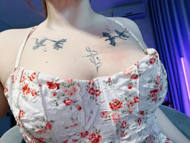 Rubbi's boobs are covered in a white, floral, lace-up dress with red roses. She has tattoos of black birds and a minimalist branch design on her chest.