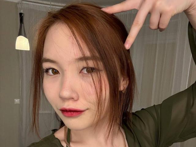 NereidaBottino with brown hair, red lips, green shirt, pointing at her head with right hand, showing off her cute face and small nose, wearing a thin necklace.