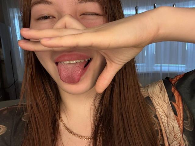 NereidaBottino with long brown hair, winking, and sticking out her tongue with her hand covering her nose, wearing a gold chain and a patterned shirt.