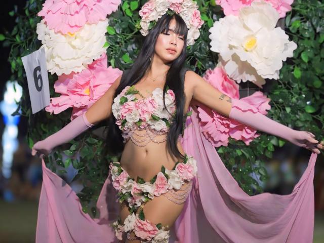 VanessaFleur in pink flower lingerie, holding pink fabric, wearing pink gloves, floral crown, and pink thigh-highs, with large pink and white flowers in the background.