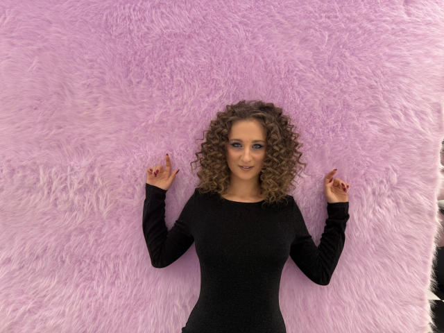 MichelleHotX69 with curly brown hair, black long-sleeve dress, standing against pink fluffy wall, hands up, fingers pointing, red nail polish, smiling, looking at camera.