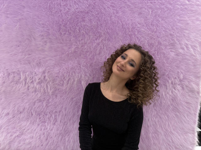 MichelleHotX69 with curly brown hair, blue eyes, and a black long-sleeve shirt, standing against a fluffy purple wall, smiling softly. Her tits are small, her ass is unseen.