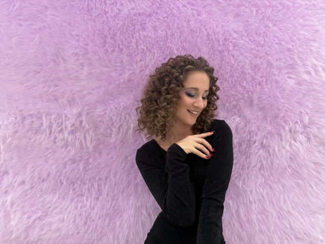 MichelleHotX69 with curly brown hair, black long-sleeve top, smiling, eyes closed, pink nail polish, hands near chest, standing against fluffy pink background.