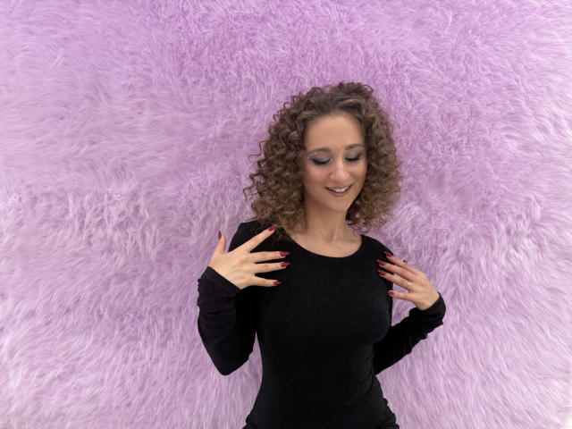 MichelleHotX69 with curly brown hair, black long-sleeve top, and red nails, smiling, hands on chest, standing against a fluffy pink wall.