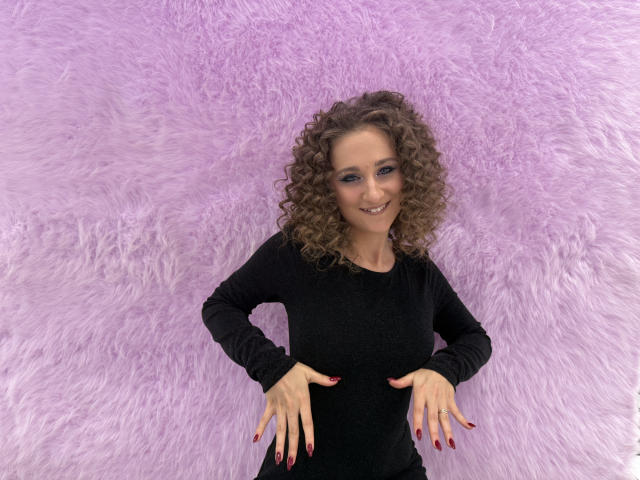 MichelleHotX69 with curly brown hair, smiling, wearing a black long-sleeve shirt, hands on hips, pink manicured nails, standing against a fluffy pink wall.