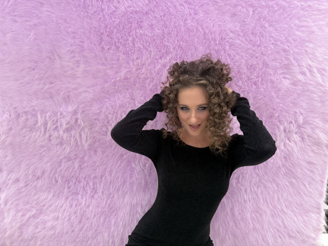 MichelleHotX69 with curly brown hair, wearing a black long-sleeve shirt, hands in her hair, smiling, standing in front of a pink, fluffy textured wall.