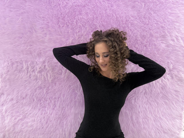 MichelleHotX69 with curly brown hair, wearing a black long-sleeve top, arms raised, looking down, smiling, standing against a purple fuzzy wall.