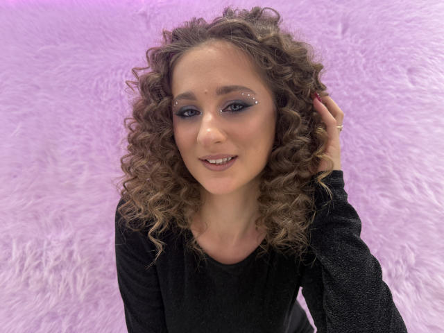 MichelleHotX69 with curly brown hair, wearing a black long-sleeve top, smiling, eyeliner with sparkling accents, hand touching her hair, purple fluffy background.