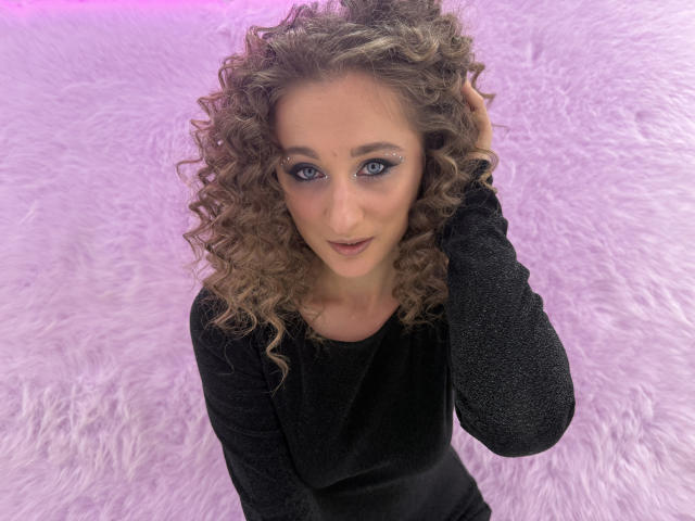 MichelleHotX69 with curly brown hair, blue eyes, wearing a black glittery long-sleeve top, looking up with a slight smile, hand in hair, purple background.