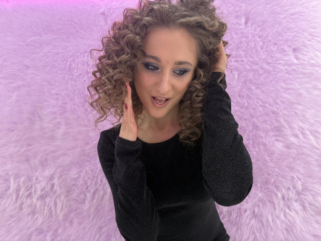 MichelleHotX69 with curly brown hair, blue eye makeup, black long-sleeve top, hands in hair, smiling, purple pink background, fluffy texture. She's looking cute, sexy, and playful.