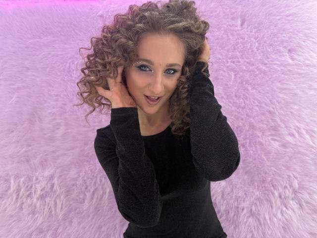 MichelleHotX69 has curly brown hair, blue eyes, and is wearing a black long-sleeve shirt, smiling with hands in her hair, standing on a pink furry surface.