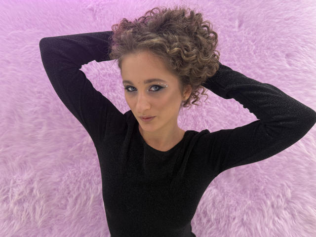 MichelleHotX69 with curly brown hair, wearing a black long-sleeve top, arms raised, hands behind head, purple fluffy background, small eye piercing, slight smile, looking up.