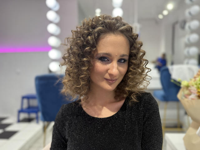 MichelleHotX69 with curly brown hair, wearing a black sparkly top, smiling slightly, blue eyes, subtle makeup, and a cute nose, in a bright indoor setting.