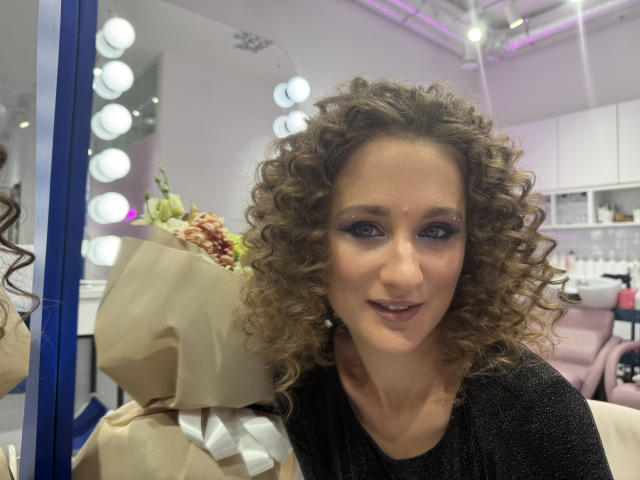 MichelleHotX69 with curly brown hair, black sparkly top, holding a brown bouquet, smiling, blue eye makeup, mirror with round bulbs, white ceiling, pink chair in background.
