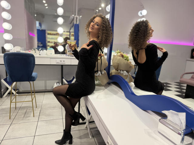 MichelleHotX69 with curly brown hair, black dress, black heels, and black stockings, sitting on a white counter, holding a drink, mirror reflection showing her ass.
