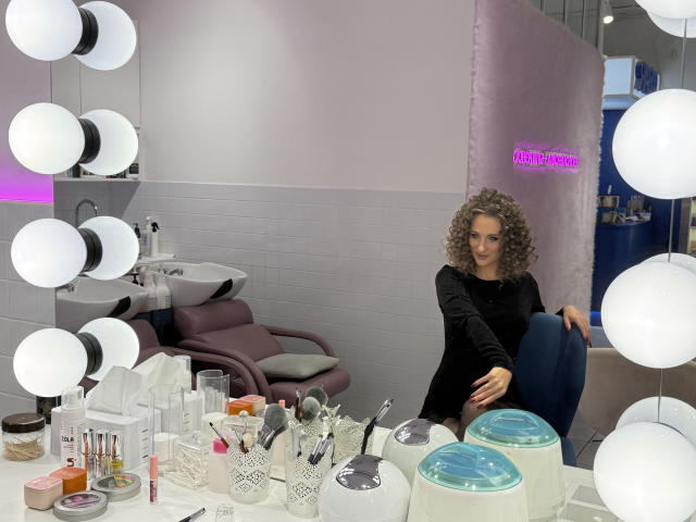 MichelleHotX69 with curly hair, black long-sleeve shirt, sitting in makeup chair, surrounded by white round lights, makeup tools, and blue-lidded containers on a white counter.