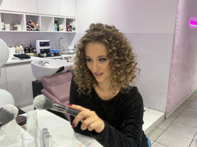 MichelleHotX69 with curly brown hair, wearing a black sweater, applying red lipstick with a brush, sitting at a makeup table, white sink, and various makeup products in the background.