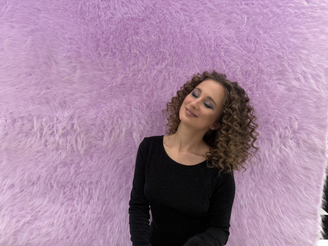 MichelleHotX69 with curly brown hair, black long-sleeve shirt, and closed eyes, standing against a fluffy pink wall, smiling slightly, looking relaxed and happy.