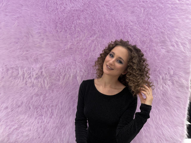 MichelleHotX69 with curly brown hair, wearing a black long-sleeve top, standing against a fluffy purple wall, smiling, and touching her hair with her right hand.