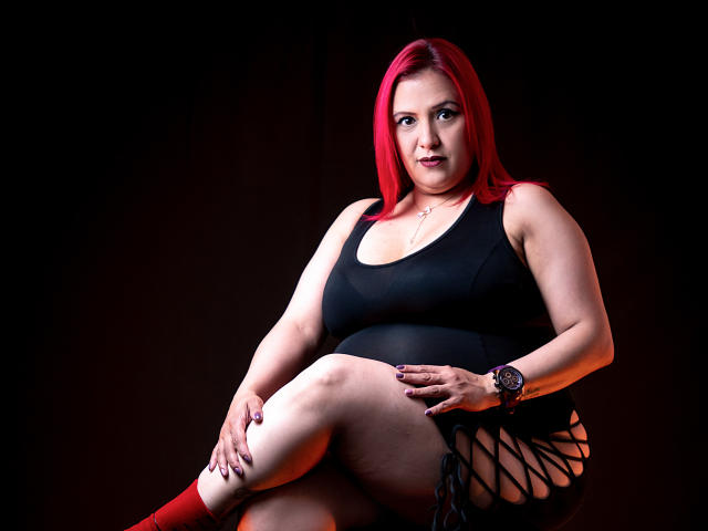 SammyOwens has red hair, wearing a black tank top and red socks, sitting with legs crossed, showing black side-tie shorts, hands on knees, and a watch on her wrist.
