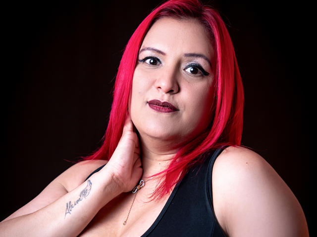 SammyOwens with bright red hair, dark eye makeup, black sleeveless top, touching her neck, showing a tattoo on her right wrist, against a dark background.