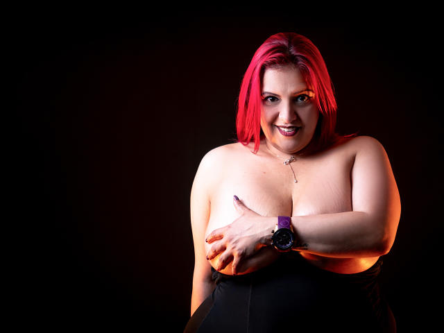 SammyOwens with red hair, smiling, topless, covering her tits with one hand, wearing a black skirt and purple watch, against a dark background.