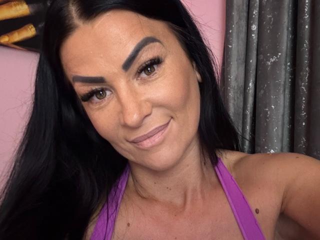 AndraD with dark hair, thick eyebrows, and purple halter top, smiling, showing small moles on her neck, and brown eyes. She's got a cute, slightly wrinkled face.
