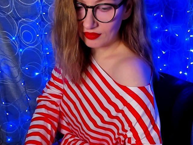 PaulinaSunny with red and white striped shirt, glasses, and red lipstick, her shoulder exposed, blue light background, and fairy lights. Her ass is not visible.