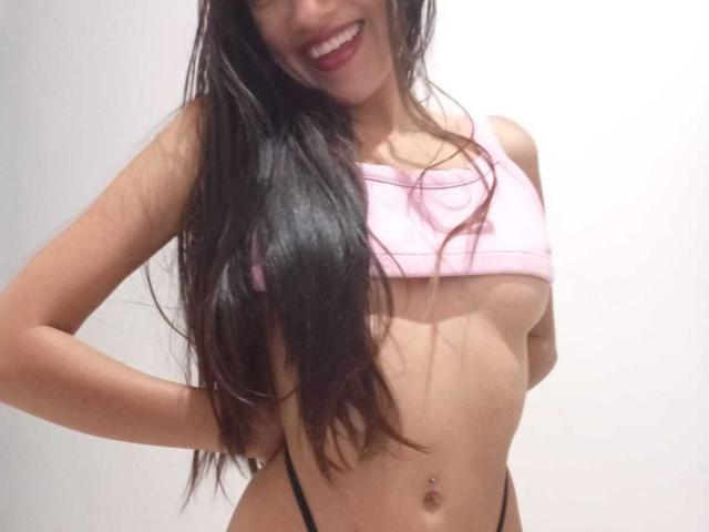 KiriamGomez with long black hair, smiling, wearing a pink crop top exposing her small tits and black thong, hands on her hips, showing a belly button piercing.