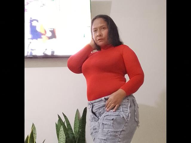 HannaPasion stands with one hand on her head, wearing a red long-sleeve top and ripped gray jeans, showing her curvy figure and small, firm ass.