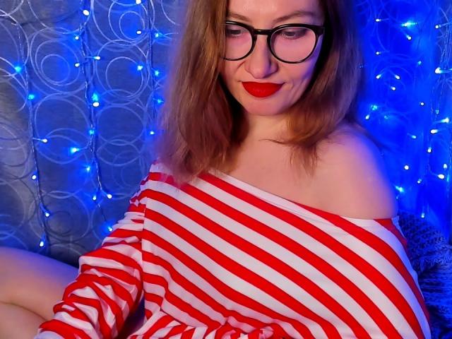 PaulinaSunny with red-striped off-shoulder top, black glasses, red lipstick, and blue lights in background, sitting with legs crossed, showing her bare shoulders and upper chest.