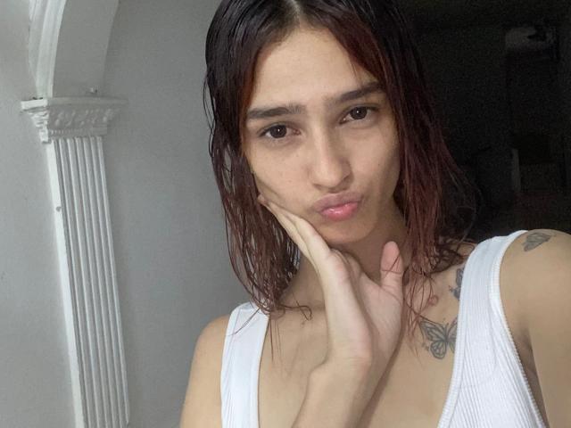 MelodyMansson with wet brown hair, pouty lips, white tank top, hand on cheek, butterfly tattoo on right shoulder, small tits, brown eyes, light skin, soft natural light.
