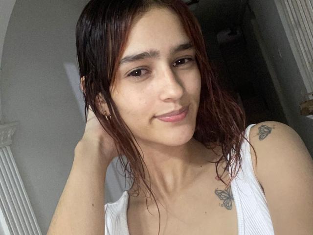 MelodyMansson has wet brown hair, a small butterfly tattoo on her right shoulder, and a butterfly tattoo on her left chest. She's wearing a white tank top.