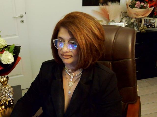 ClassybutNaughty with red hair, wearing clear glasses reflecting light, black blazer, and multiple necklaces, sitting in a brown leather chair, smiling. Boobs partially visible.