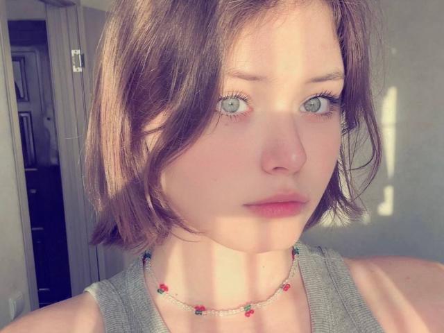 NikolKitt has light green eyes, a small nose, and short brown hair. She's wearing a grey tank top and a beaded necklace with red and green flowers.
