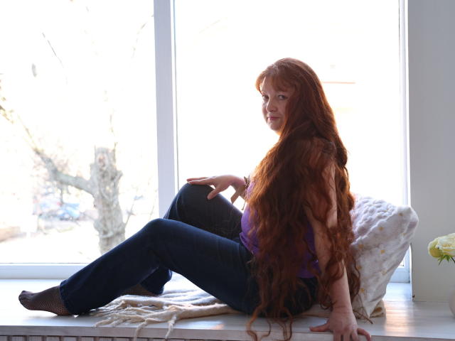 OVanessaHotly with long red hair, purple top, blue jeans, sitting on window ledge, legs crossed, white pillow behind, sunlight, relaxed, slightly smiling, bare feet, white wall.