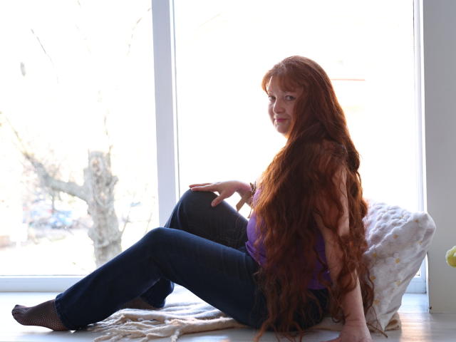 OVanessaHotly with long red hair, purple top, blue jeans, and black socks, sitting on a window sill, smiling, legs crossed, white pillow behind her.