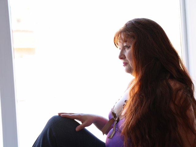 OVanessaHotly with long red hair, purple top, and blue jeans, sitting in bright light, looking down with a pensive expression, hands resting on her knee.