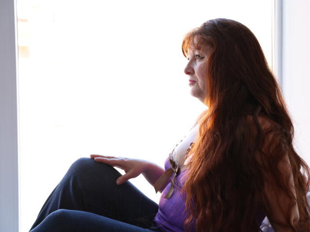 OVanessaHotly with long red hair, sitting, wearing a purple top and blue jeans, sunlight in profile, hands on knee, looking right, soft light, relaxed, casual, natural.