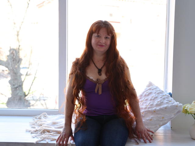 OVanessaHotly has long red hair, wearing a purple top and blue jeans, sitting on a windowsill with a white pillow and beige blanket, smiling with natural light.