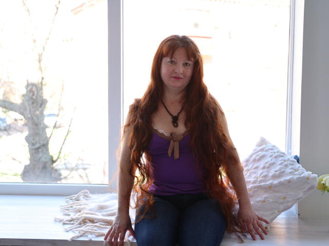 OVanessaHotly with long red hair, purple top, and blue jeans, sitting on a windowsill, hands on her thighs, looking at the camera, white pillow behind her.