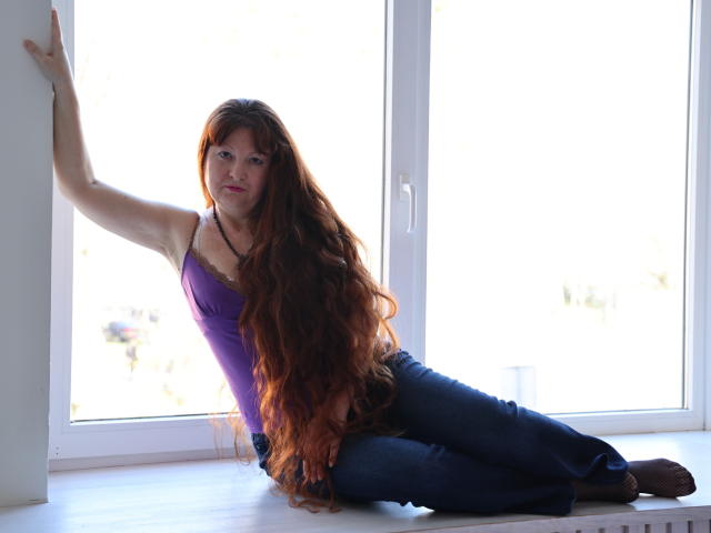 OVanessaHotly with long red curly hair, purple top, and blue jeans, sitting on window sill, arm raised, white wall behind, bright sunlight, sexy, relaxed, attractive.