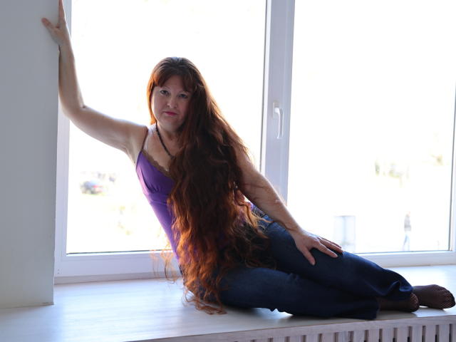 OVanessaHotly with long red hair, purple tank top, and blue jeans, leaning on a window, hand on hip, looking at camera, sunlight behind her.