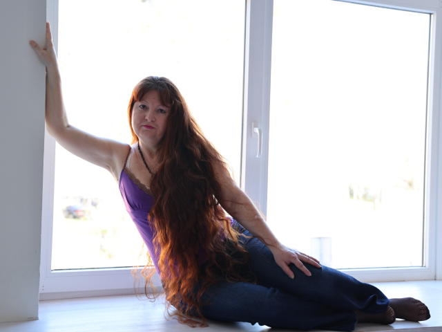 OVanessaHotly with long red hair, purple top, and blue jeans, sitting on white windowsill, leaning on wall, looking up, hand on thigh, sunlight behind her.