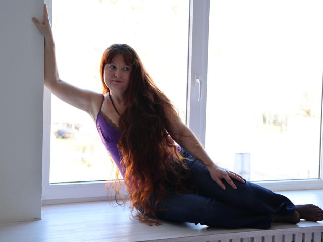 OVanessaHotly with long red hair, purple tank top, and blue jeans, sitting on window ledge, hand on wall, other hand on knee, looking at camera, smiling.