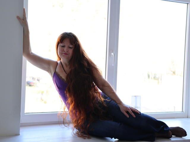 OVanessaHotly with long red hair, purple top, and blue jeans, leaning against a window, her hand on her thigh, and her other arm on the window frame.
